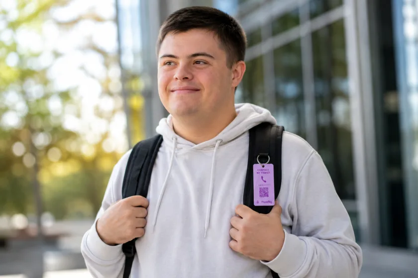 A young man wearing a backpack with a lavender PhoneTag contact tag clipped to the strap